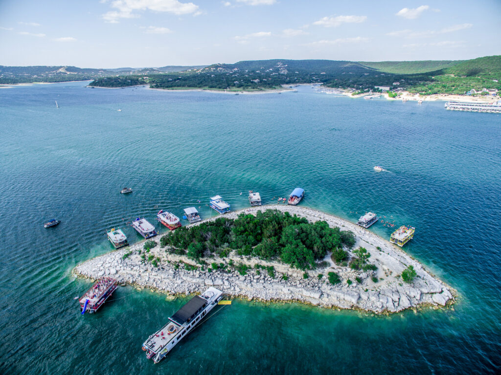 Aerial view of a Lake Travis cove with a dock and several boats moored around a small rocky island, the kind of waterfront a Hill Country dock builder works on.